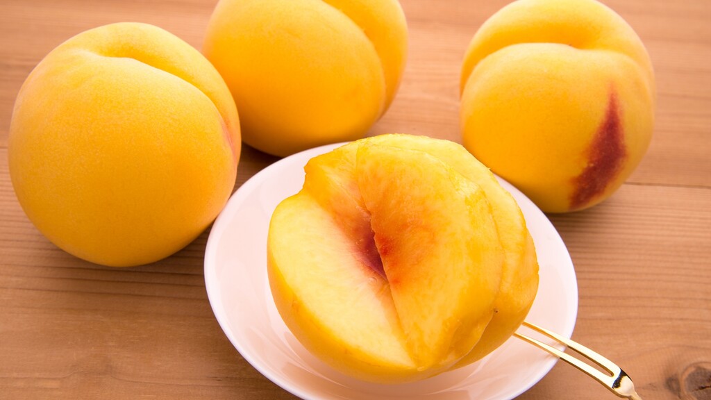Sliced yellow peach on a white plate with whole peaches in the background.