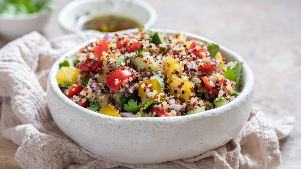 Quinoa salad with tomatoes, cucumber, bell peppers, herbs, and dressing in a white ceramic bowl.