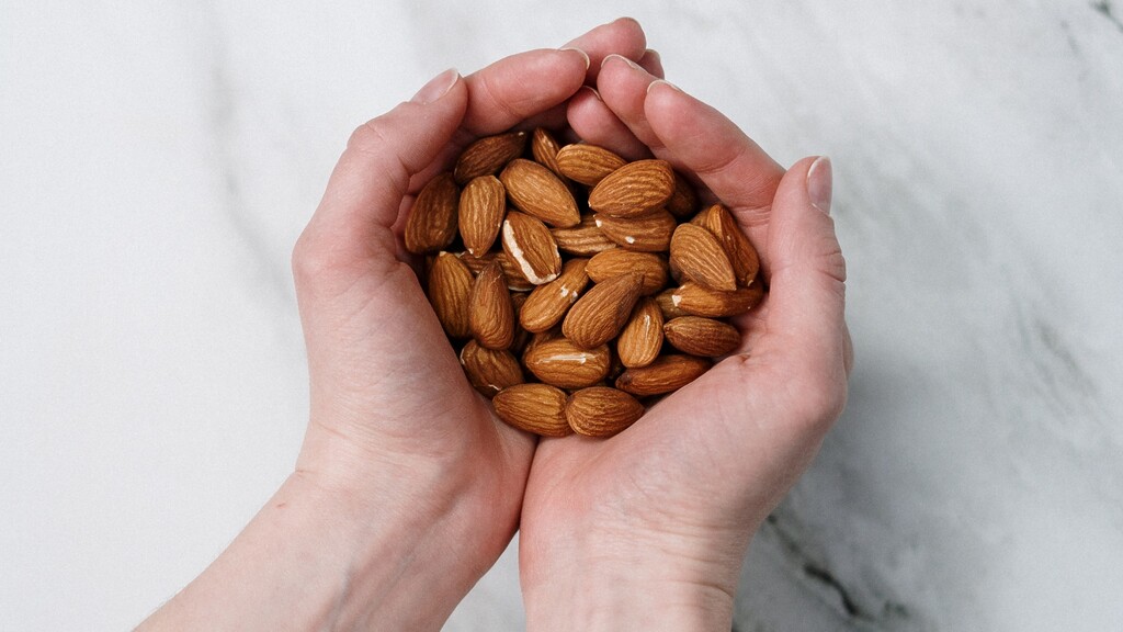 Hands holding a handful of raw almonds on a light marble countertop.