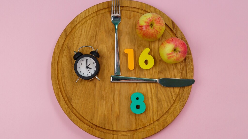 16:8 intermittent fasting concept with a clock, fork, knife, apples, and 16 and 8 numbers arranged on a wooden plate.