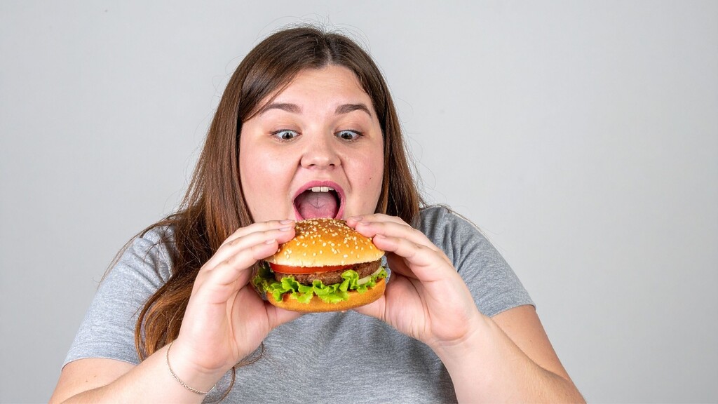 A person in a gray shirt holds a large burger close to their mouth against a plain light background.
