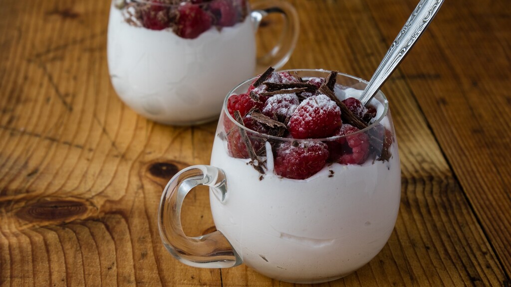 Frozen raspberries with cream and dark chocolate shavings served in small glass cups on a wooden table.