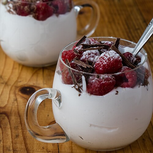 Frozen raspberries with cream and dark chocolate shavings served in small glass cups on a wooden table.
