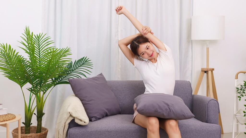 Woman doing a gentle seated side stretch on a sofa in a bright living room.