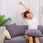 Woman doing a gentle seated side stretch on a sofa in a bright living room.