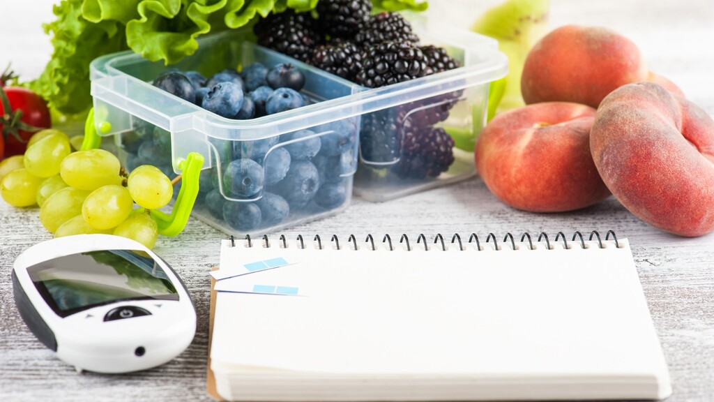 Blood glucose meter and blank notebook beside fresh berries, grapes, peaches, and leafy greens for diabetes meal tracking.