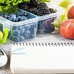 Blood glucose meter and blank notebook beside fresh berries, grapes, peaches, and leafy greens for diabetes meal tracking.