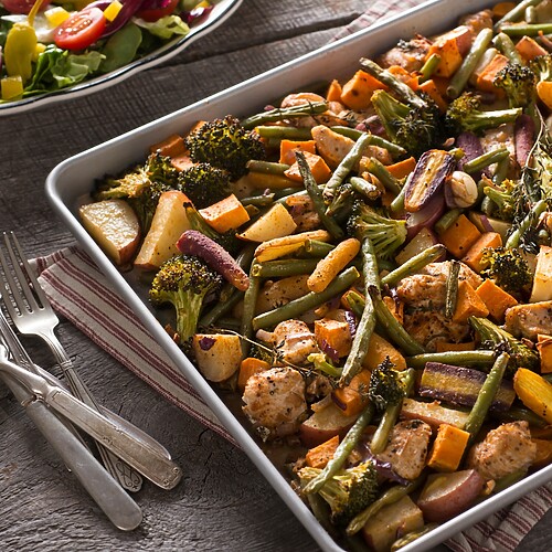 Sheet pan dinner with roasted chicken, broccoli, green beans, onions, and sweet potatoes on a rustic wooden table.