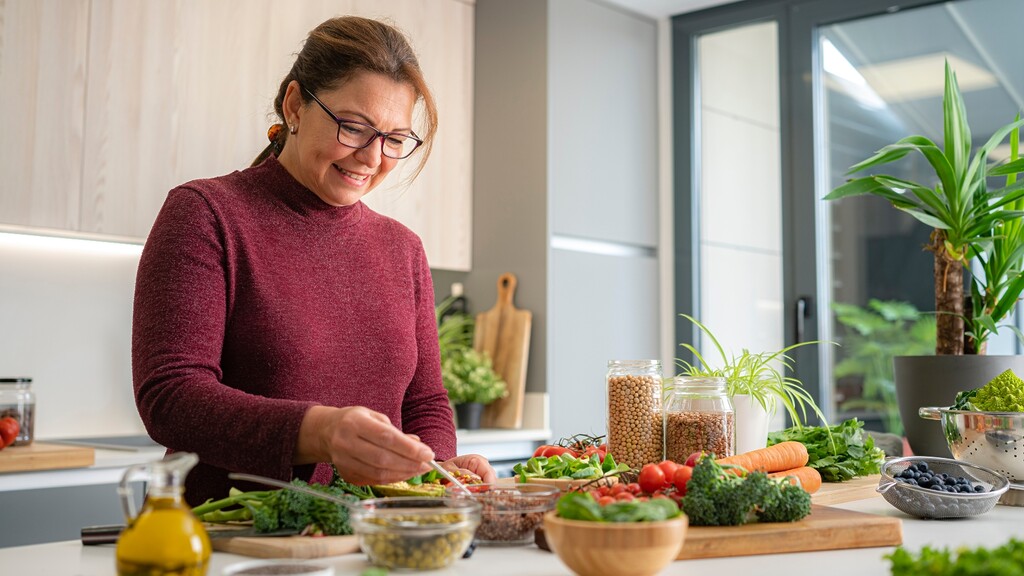 Woman prepares a balanced meal with vegetables, legumes, olive oil, and berries in a bright kitchen.