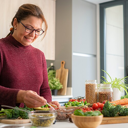 Woman prepares a balanced meal with vegetables, legumes, olive oil, and berries in a bright kitchen.