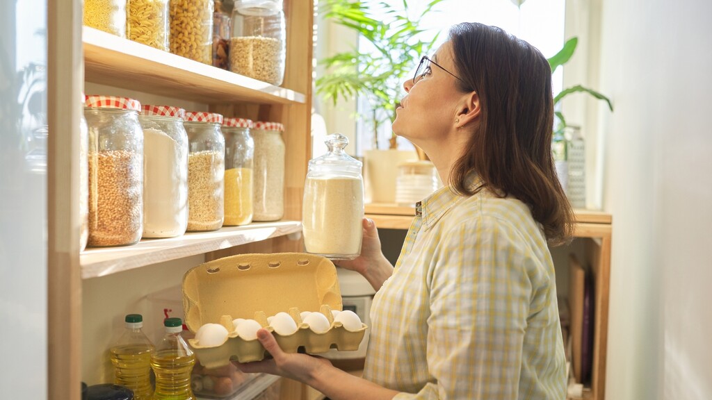 Woman organizing pantry shelves with glass jars, eggs, and cooking oils in a bright kitchen.