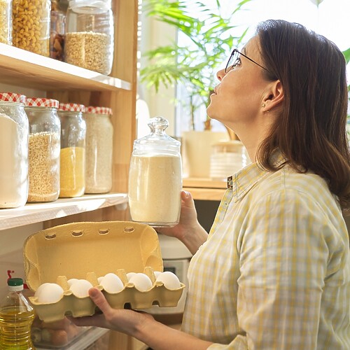 Woman organizing pantry shelves with glass jars, eggs, and cooking oils in a bright kitchen.