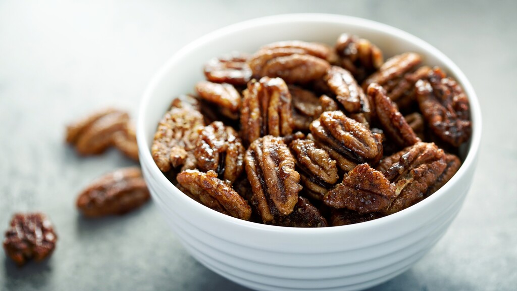 Bowl of cinnamon-dusted pecans on a light surface, with a few pecans scattered beside it.