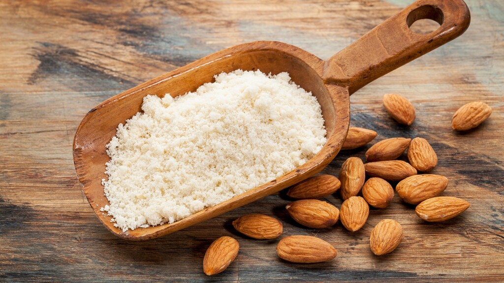 Almond flour in a wooden scoop with whole almonds on a rustic wooden table.