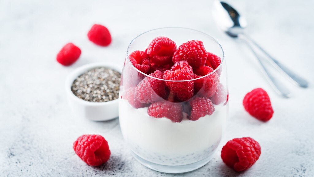 Greek yogurt topped with fresh raspberries, with chia seeds and a spoon on a light kitchen surface.
