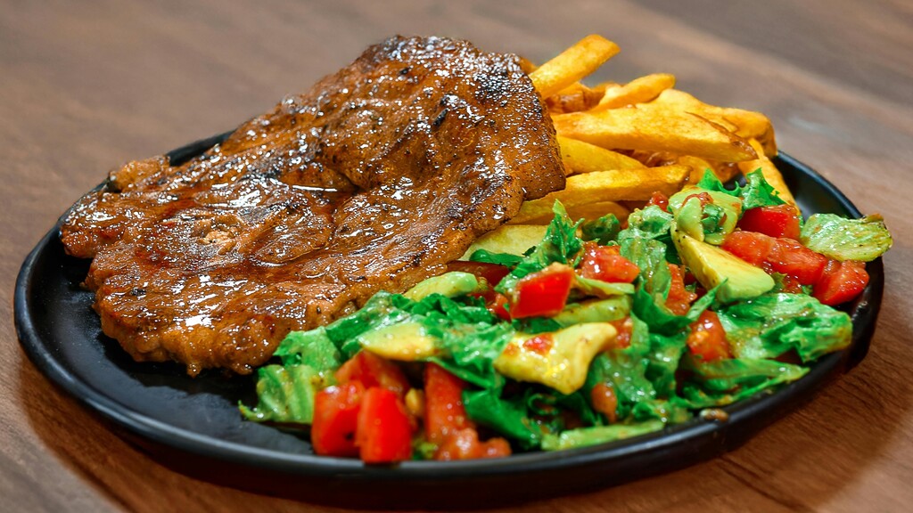 A plate of glazed grilled meat served with fries and a fresh vegetable salad on a wooden table.