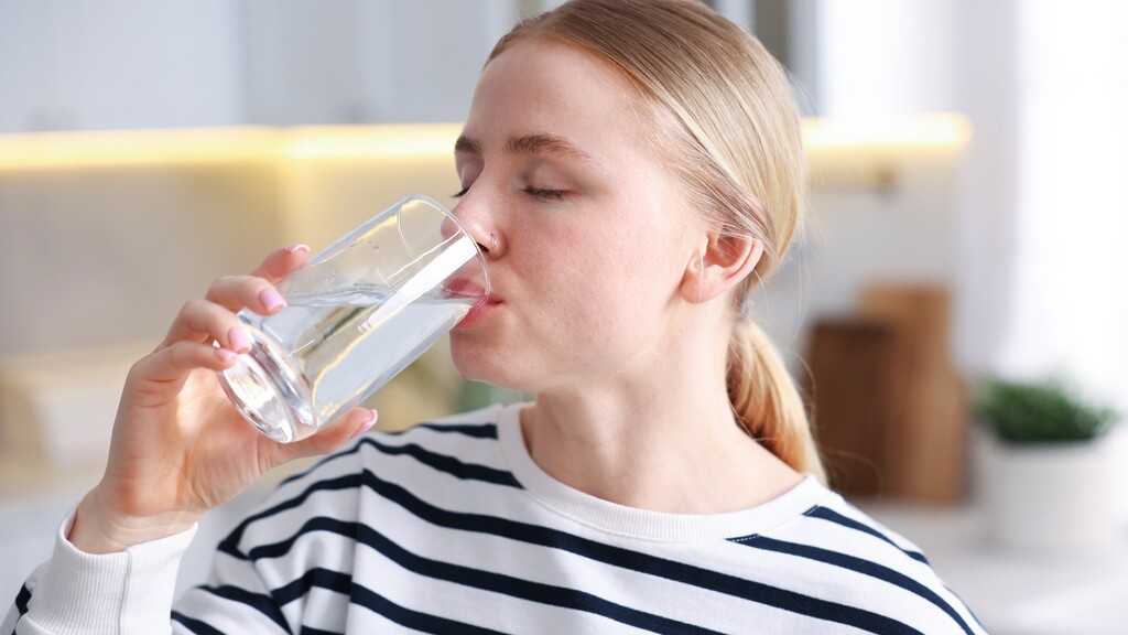 Woman drinking a glass of water in a bright kitchen.