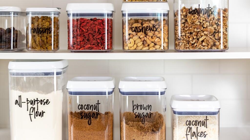 Organized pantry shelf with clear containers of nuts, raisins, flour, coconut sugar, brown sugar, and coconut flakes.