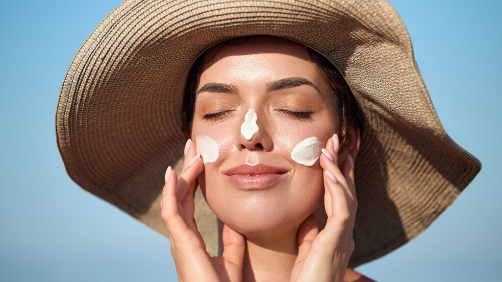 Woman applying sunscreen while wearing a sun hat to protect her face from UV damage.