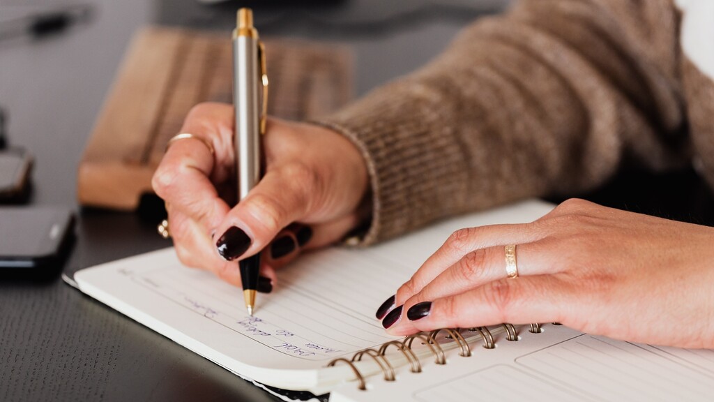 Close-up of hands writing notes in a spiral notebook with a pen.