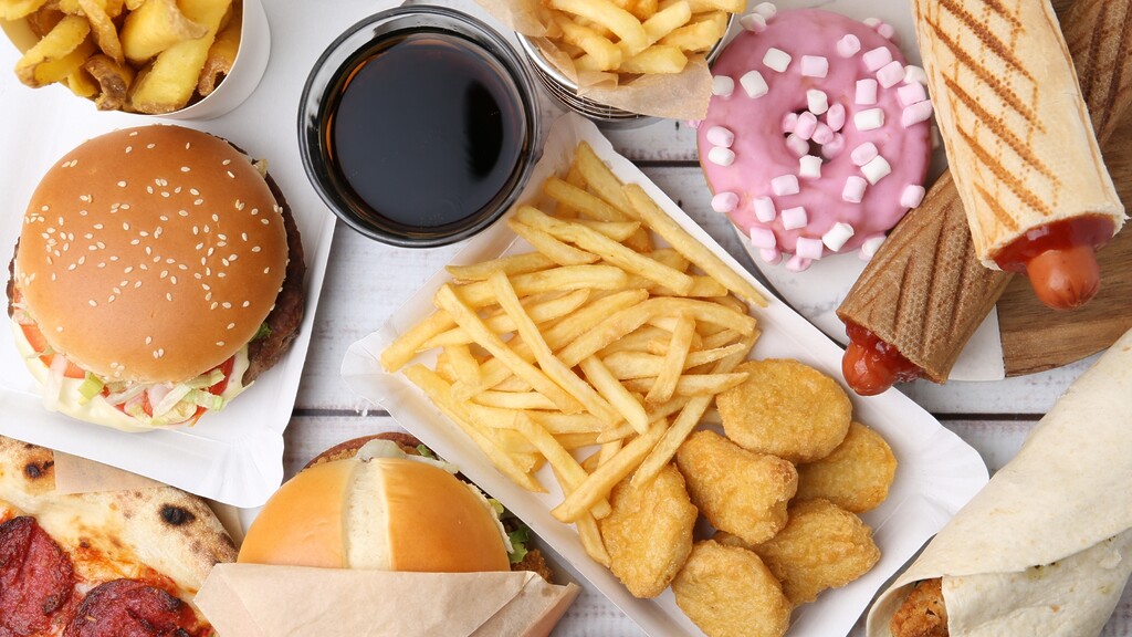 Overhead spread of fast food including burgers, fries, chicken nuggets, soda, pizza, hot dogs, wraps, and a pink doughnut.