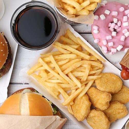 Overhead spread of fast food including burgers, fries, chicken nuggets, soda, pizza, hot dogs, wraps, and a pink doughnut.