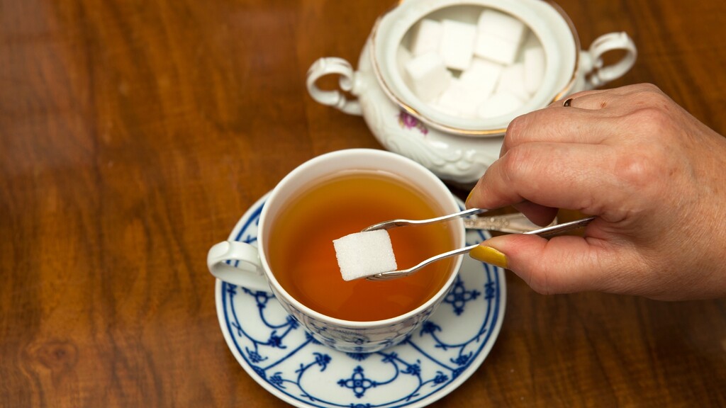 Hand placing a sugar cube into a cup of tea beside a sugar bowl on a wooden table.