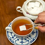 Hand placing a sugar cube into a cup of tea beside a sugar bowl on a wooden table.