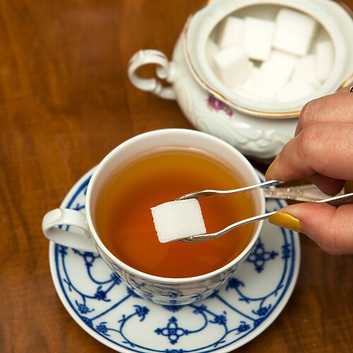 Hand placing a sugar cube into a cup of tea beside a sugar bowl on a wooden table.