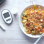 Blood glucose meter beside a bowl of vegetables and protein, representing diabetes-friendly meal planning.