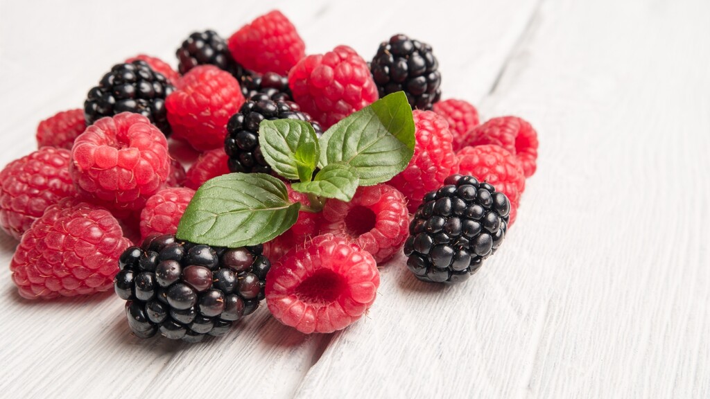 Fresh raspberries and blackberries with mint leaves on a white wooden surface.
