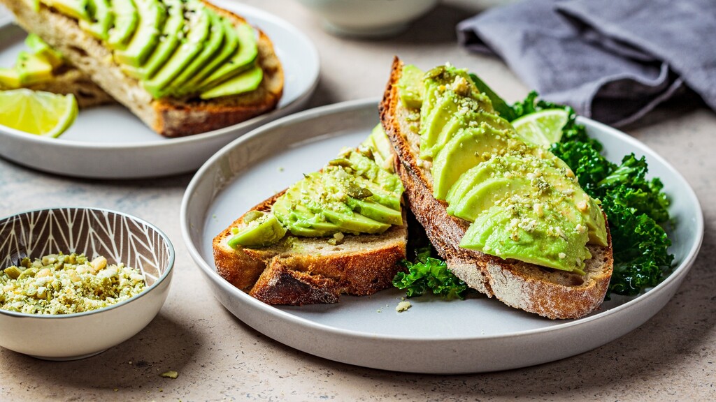 Avocado toast served with kale, lime wedges, and seeds on a plate.