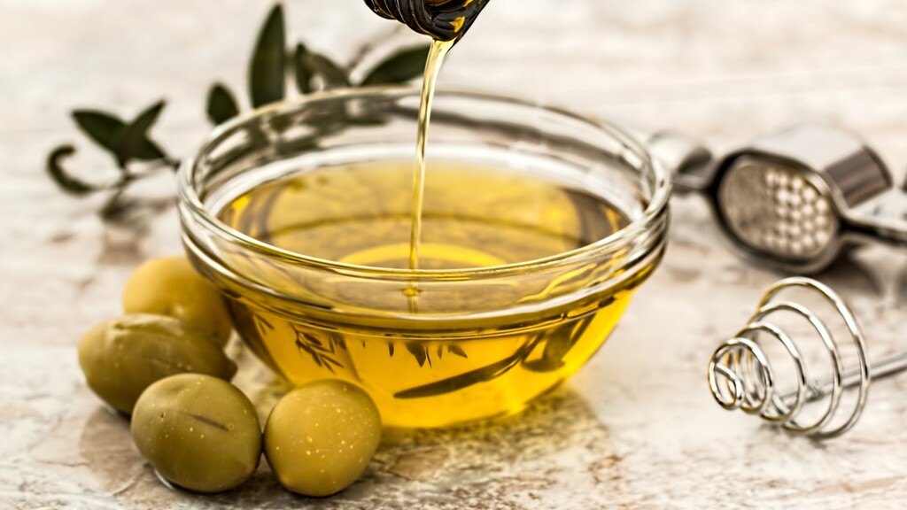Extra virgin olive oil being poured into a glass bowl with green olives nearby.