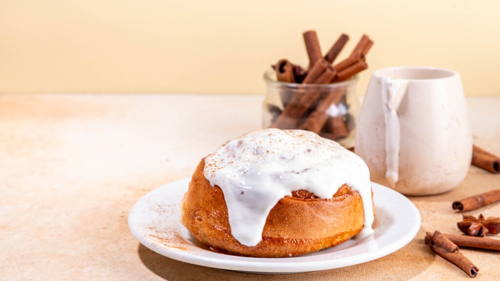 Single cinnamon roll with cream cheese frosting on a white plate, with cinnamon sticks in the background.