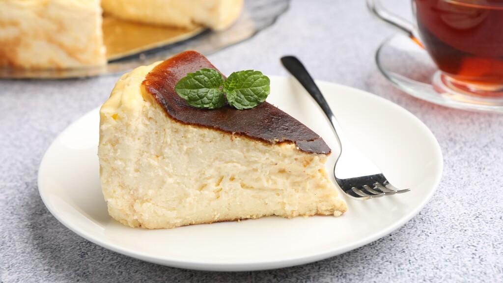 Slice of creamy baked cheesecake on a white plate, topped with mint and served with tea.