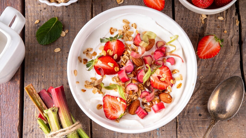 Greek yogurt topped with strawberries, almonds, rhubarb, and granola on a wooden table.