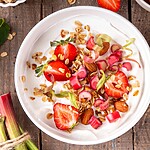 Greek yogurt topped with strawberries, almonds, rhubarb, and granola on a wooden table.