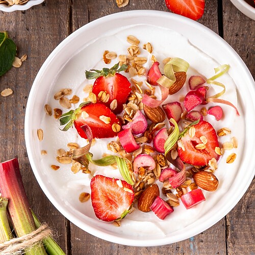Greek yogurt topped with strawberries, almonds, rhubarb, and granola on a wooden table.