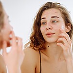 Young woman examining acne-prone skin in a bathroom mirror.