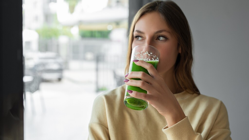 Woman drinking a green smoothie while looking out a window in a bright indoor setting.