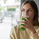 Woman drinking a green smoothie while looking out a window in a bright indoor setting.