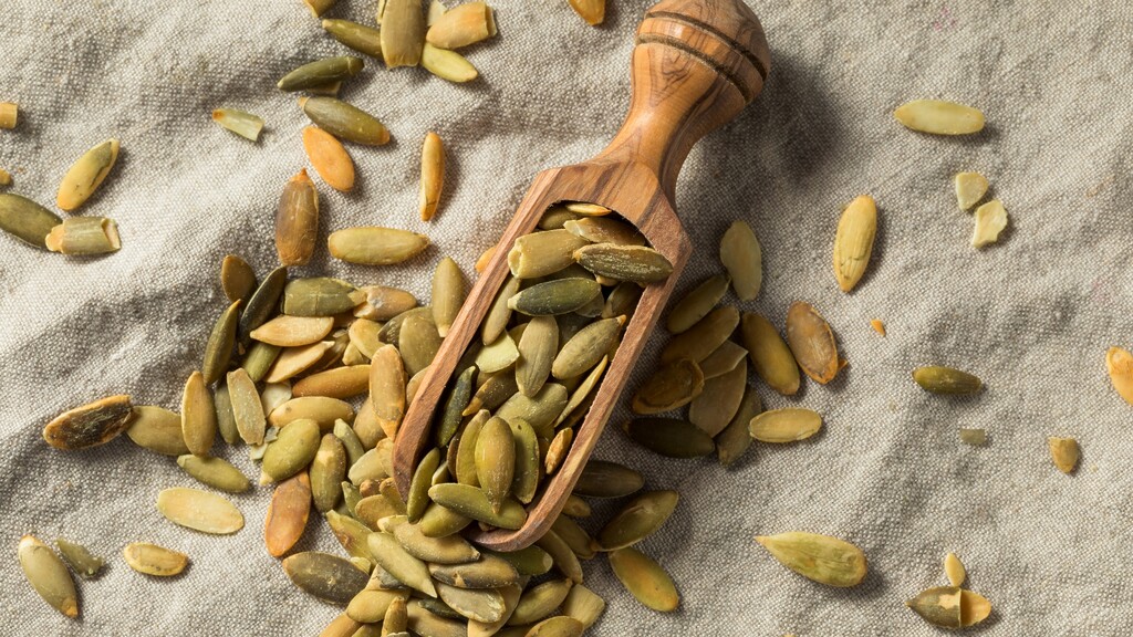 Shelled pumpkin seeds scattered around a wooden scoop on a textured linen cloth.
