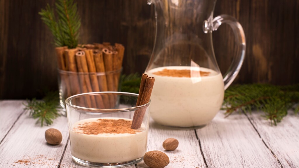 A glass of warm milk with cinnamon beside a pitcher and cinnamon sticks.