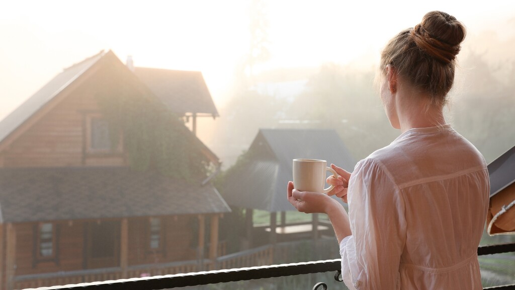 Woman holding a mug on a balcony in soft morning sunlight, representing a calm morning routine for supporting stress balance and cortisol regulation.