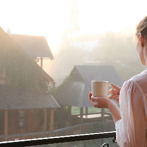 Woman holding a mug on a balcony in soft morning sunlight, representing a calm morning routine for supporting stress balance and cortisol regulation.
