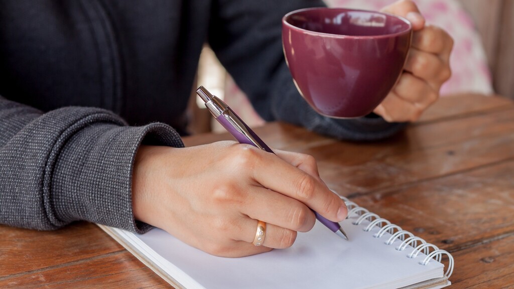 Hand writing in a notebook beside a coffee mug, a simple evening habit for clearing tomorrow’s tasks.