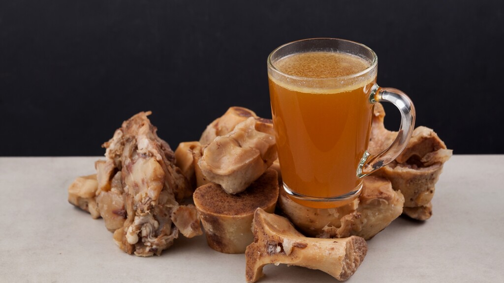 Glass mug of warm bone broth served with roasted bones on a dark kitchen background