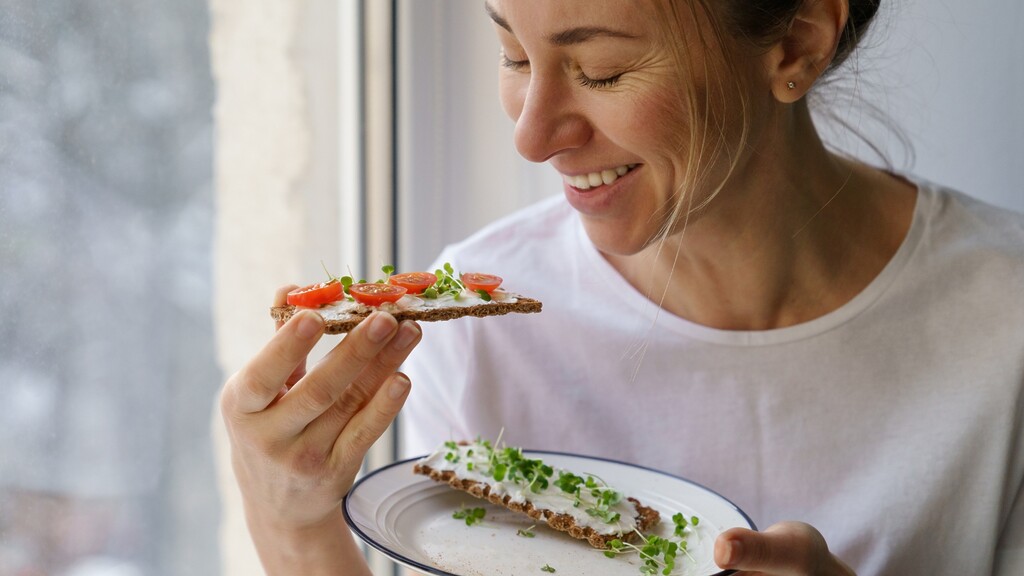 A smiling woman eating whole-grain crispbread topped with soft cheese, tomatoes, and microgreens by a window.