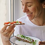 A smiling woman eating whole-grain crispbread topped with soft cheese, tomatoes, and microgreens by a window.