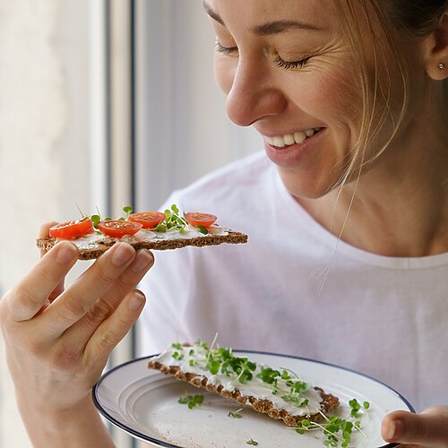 A smiling woman eating whole-grain crispbread topped with soft cheese, tomatoes, and microgreens by a window.